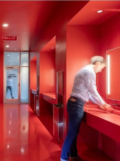 A man in a deep red all-gender bathroom washes his hands at the sink as a corridor behind him indicates open restroom stalls.